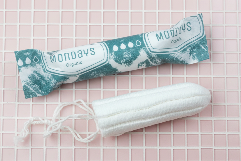 Mondays certified organic tampon non-applicator one wrapped in paper, one unwrapped. Both placed on pink mosiac tiles. 